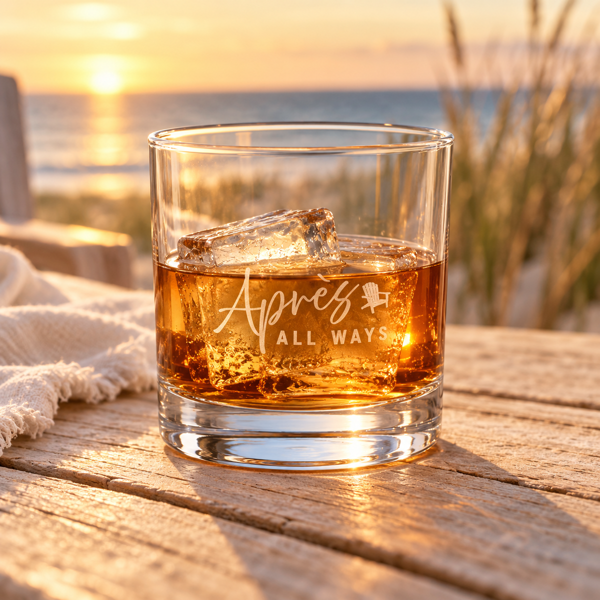 Après All Ways rocks glass with the brand logo, filled with an amber drink and ice on a wooden table overlooking the ocean at sunset.