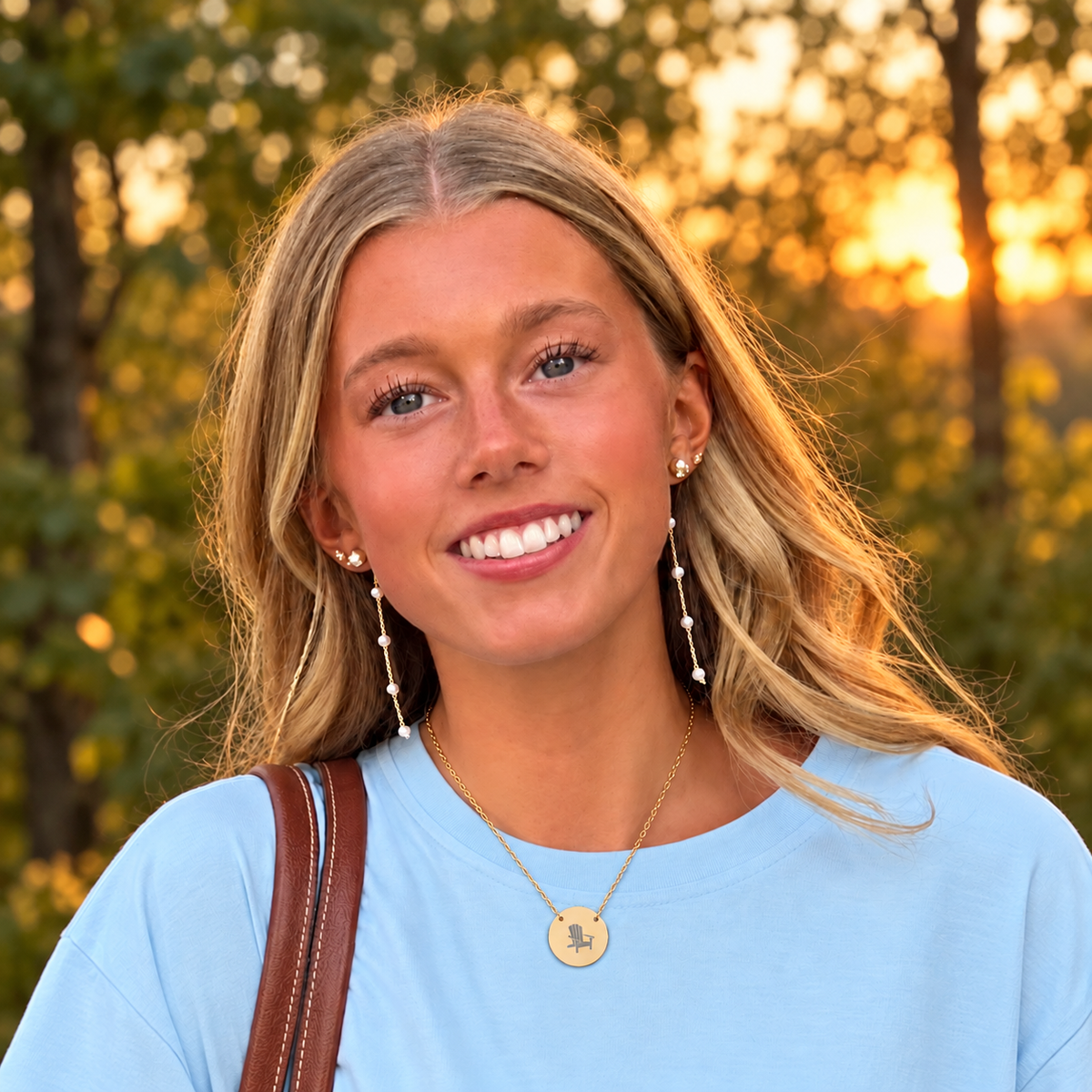 Model wearing the Après All Ways gold necklace with a round Adirondack chair engraved pendant over a light blue shirt, with trees at sunset in the background.