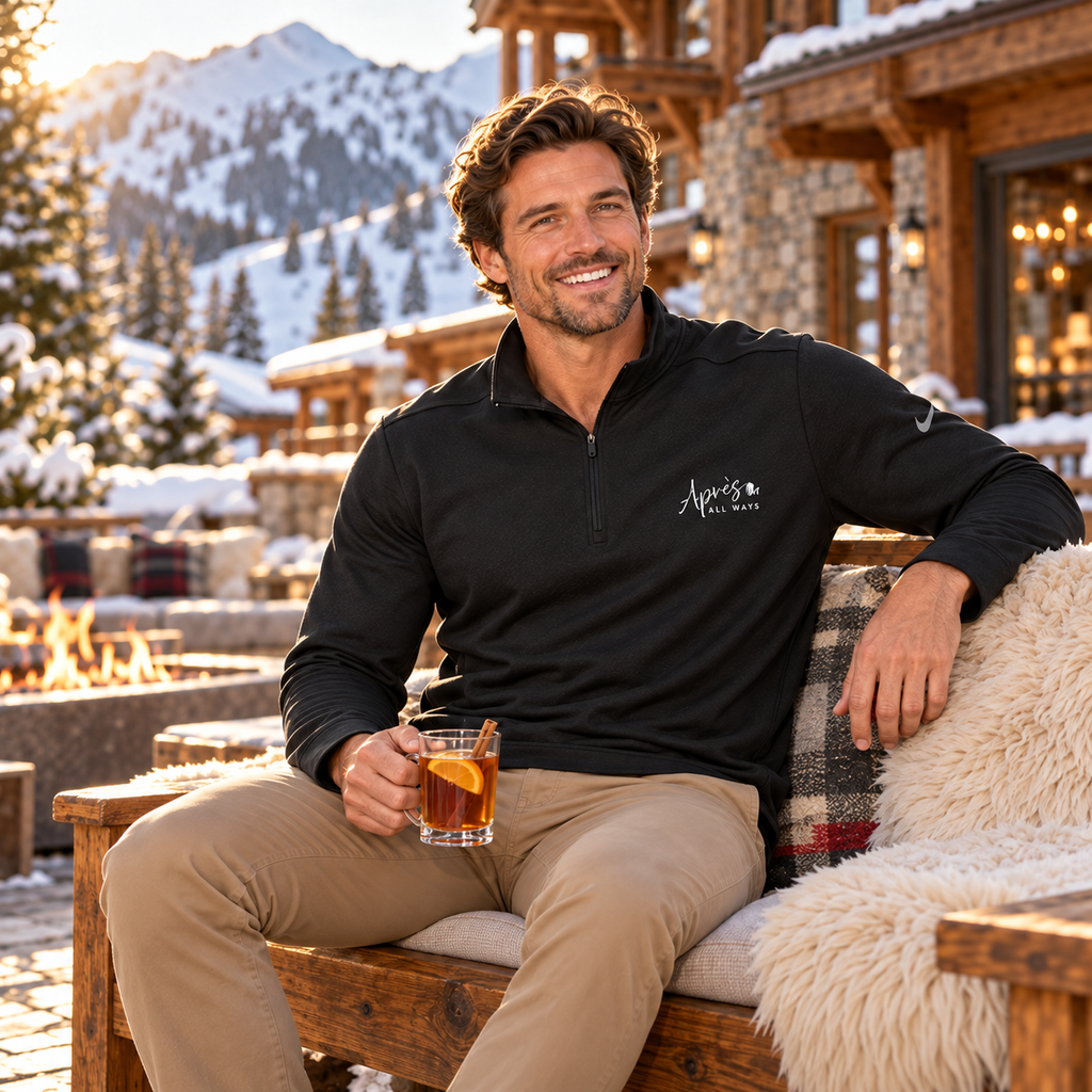 Man wearing a black Après All Ways Nike half-zip pullover, seated with a drink in a snowy ski lodge courtyard beside a fire pit.