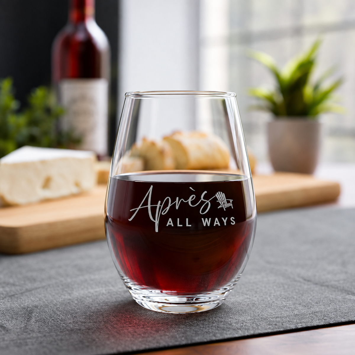 Après All Ways stemless wine glass with a white logo and Adirondack chair icon, filled with red wine beside a cheese and bread board.
