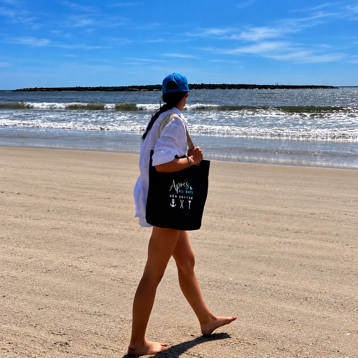 Woman in a blue cap and white shirt carrying a black Après All Ways tote with anchor, crossed skis, and golf tee icons while walking barefoot along a sandy beach.
