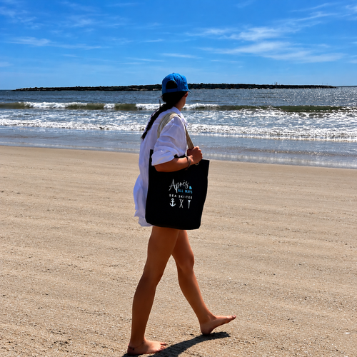 Woman in a blue cap and white shirt carrying a black Après All Ways tote with anchor, crossed skis, and golf tee icons while walking barefoot along a sandy beach.