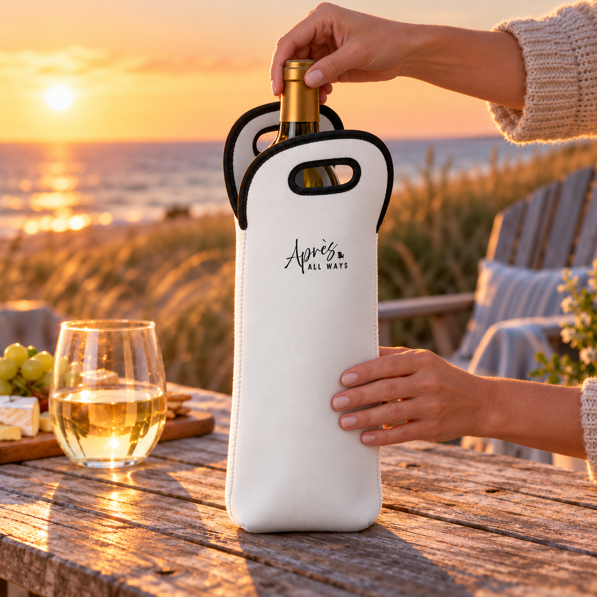 Hands lifting a wine bottle from a white Après All Ways tote with black trim, beside a wine glass and cheese board overlooking the ocean at sunset.